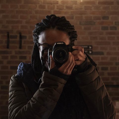 A person with braided hair and glasses takes a photo with a camera in front of a brick wall, under a bright light.