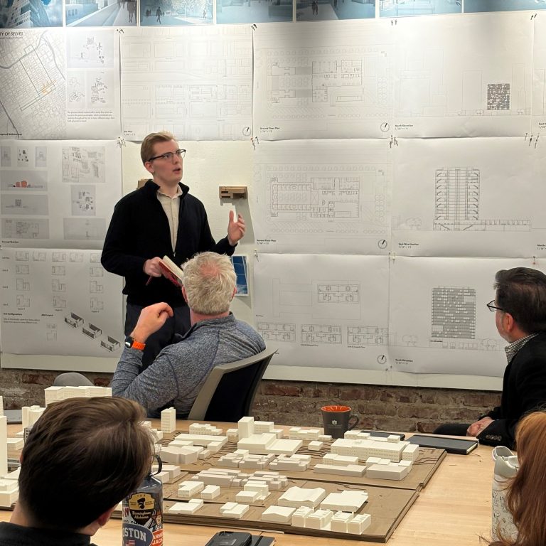 A person stands and presents architectural plans to a seated group in a studio, with design drawings and models displayed on the wall and table.
