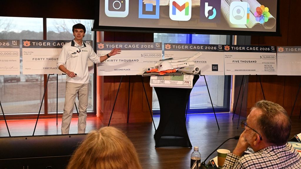 A person presents in front of judges, with a large screen showing app icons, oversized checks on display, and a model aircraft on a table at the Tiger Cage 2026 event.