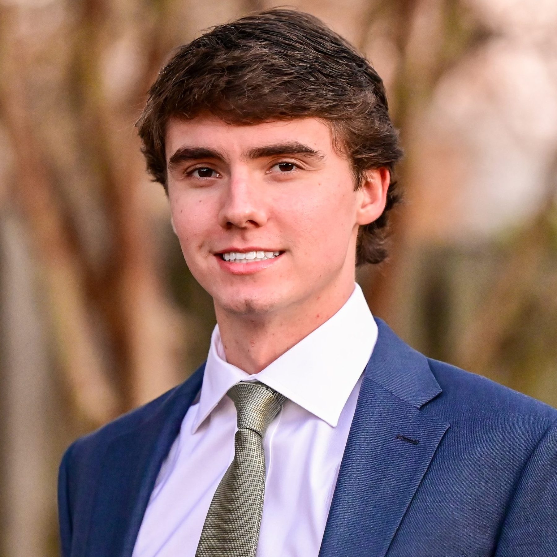 A young man in a blue suit, white shirt, and light green tie stands outdoors, smiling at the camera. Blurred trees and a fence are in the background.
