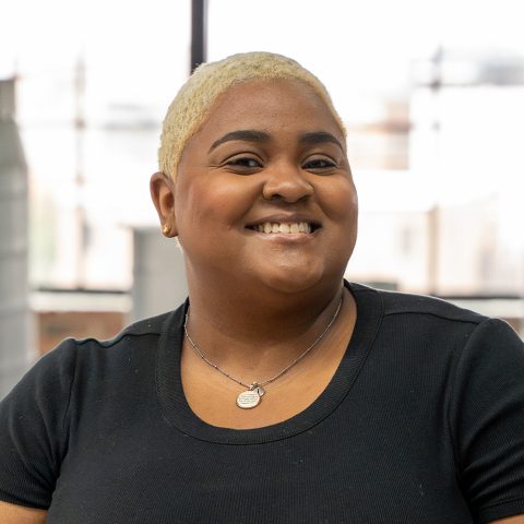 A person with short blond hair and a black shirt stands indoors, smiling, with industrial ventilation pipes and large windows in the background.