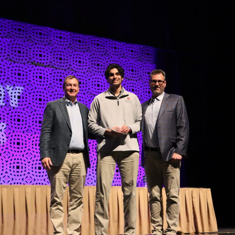 Three men stand on a stage with a patterned purple backdrop, posing for a photo. The man in the center holds an award or certificate.