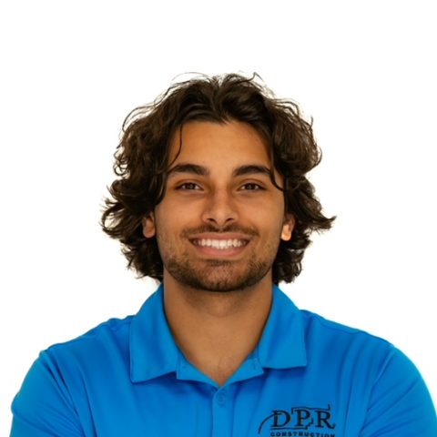 A person with wavy dark hair and a beard smiles, wearing a blue DPR Construction polo shirt, against a plain white background.