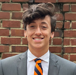 A young man in a gray suit, white shirt, and orange-and-black striped tie stands in front of a brick wall, smiling at the camera.