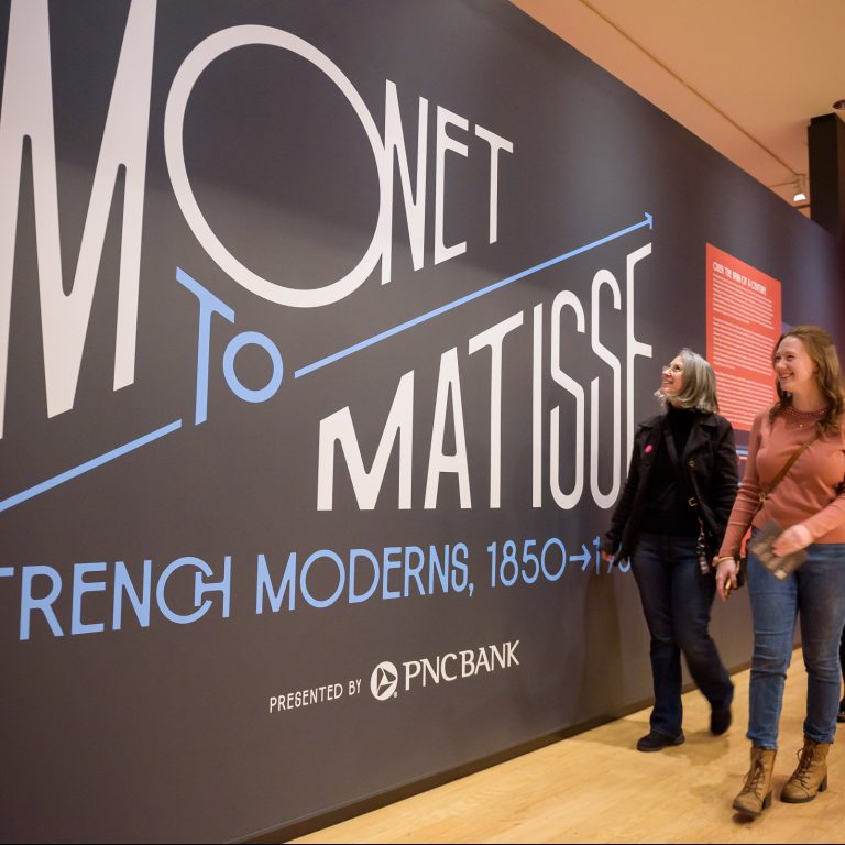 Three women walk past a large wall display reading “Monet to Matisse: French Moderns, 1850–1950” at an art exhibition presented by PNC Bank.