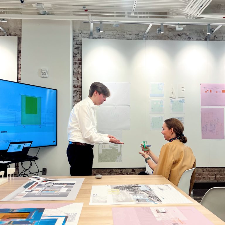 Two people discuss a small architectural model in a modern office with design plans and drawings displayed on the walls and table.