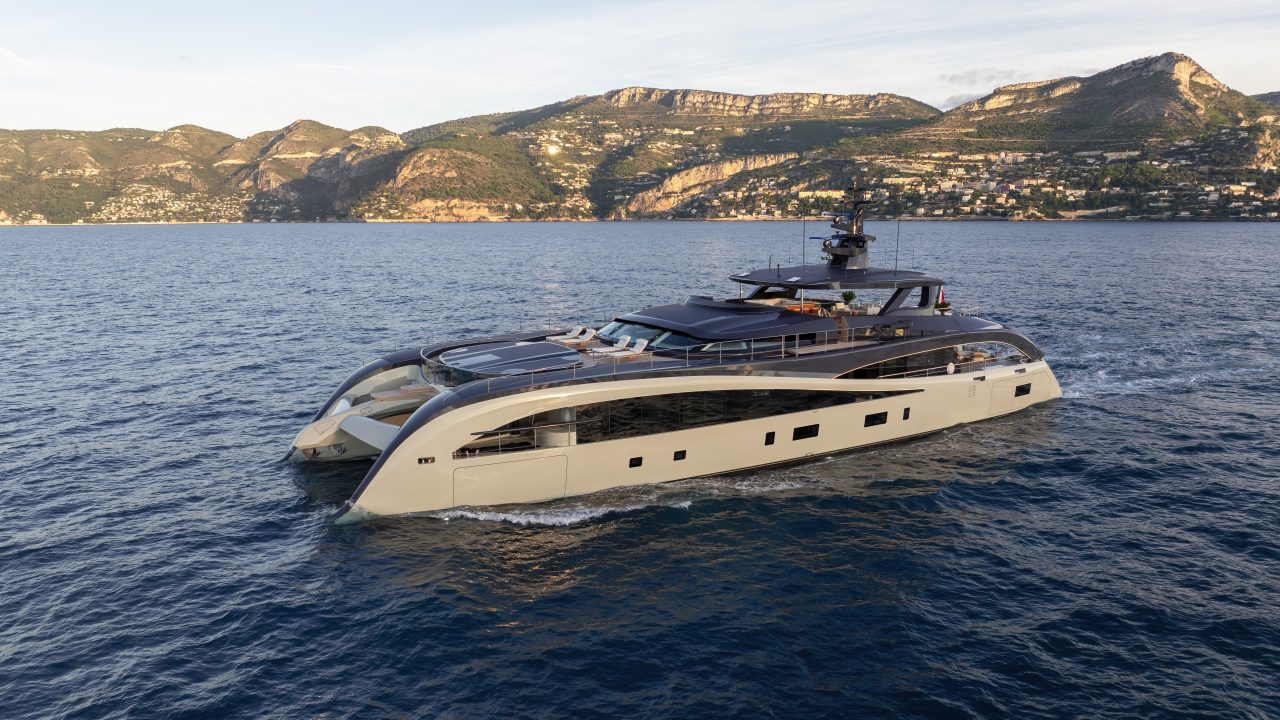 A modern luxury catamaran yacht cruises on calm blue water near a coastline with mountains and buildings in the background.