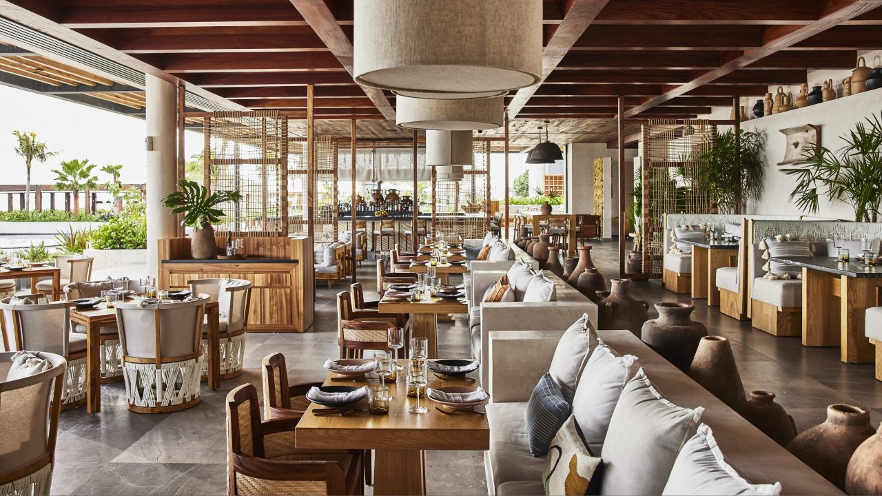 Spacious, modern restaurant with wooden furniture, neutral decor, large windows, potted plants, and neatly set tables ready for diners.