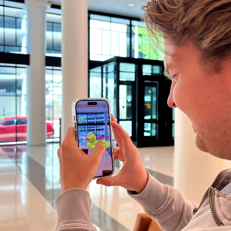 A person holds a smartphone, displaying an augmented reality yellow rubber duck, inside a modern building with large windows and white columns.