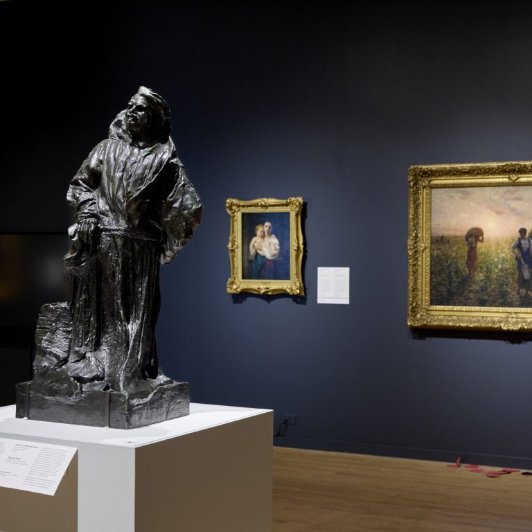 A museum gallery with a bronze statue on a pedestal in the foreground and two framed paintings on a dark wall in the background.