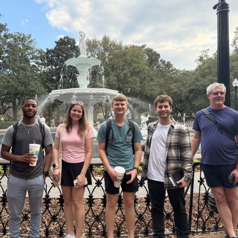 Five people standing in front of a large white fountain in a park, holding drinks and smiling, with trees and cloudy sky in the background.