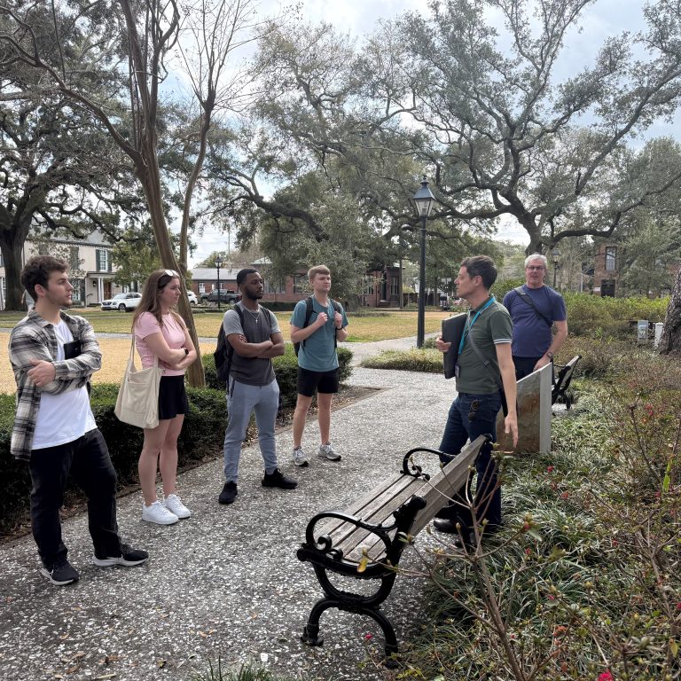 A group of six people stand and talk outdoors near a bench and trees on a paved pathway in a park-like setting.