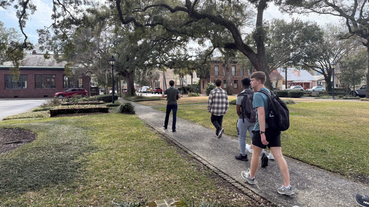 Five people walk along a sidewalk through a grassy, tree-lined park area on a cloudy day, with buildings and parked cars in the background.