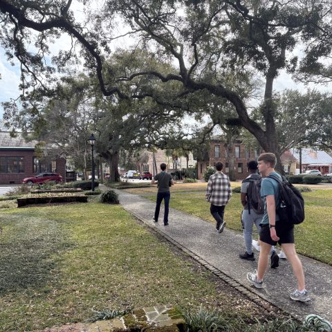 Five people walk along a sidewalk through a grassy, tree-lined park area on a cloudy day, with buildings and parked cars in the background.