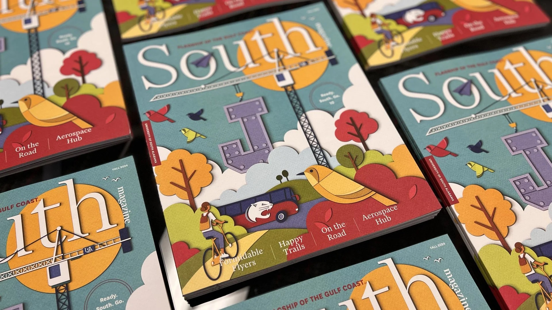 Stacks of South magazines featuring a colorful illustrated cover with trees, a crane, a cyclist, and a yellow bird; various headlines are visible on the covers.