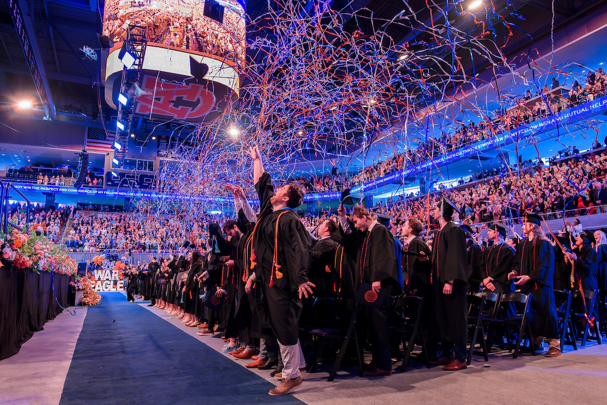 Graduates in caps and gowns celebrate at a commencement ceremony, standing as colorful streamers fall from above in a crowded indoor arena.