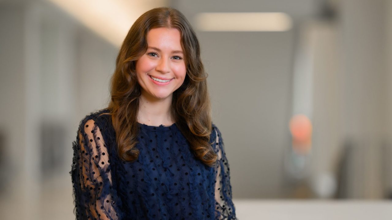 A young woman with long brown hair, wearing a navy blue textured blouse, smiles while sitting at a white table in a bright, modern indoor setting.
