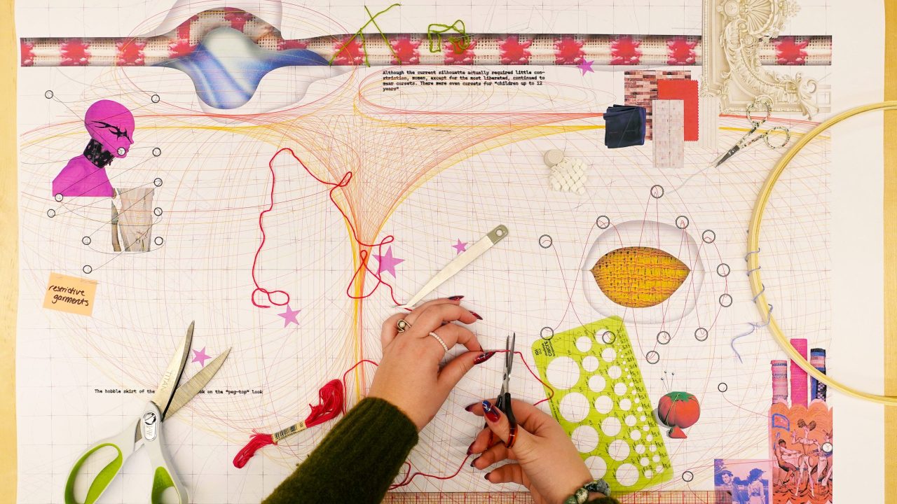 A pair of hands work with embroidery thread and tools on a colorful, mixed-media design sheet featuring drawings, patterns, and sewing materials.