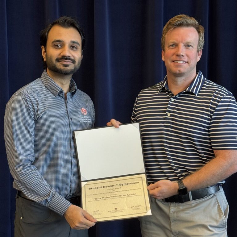 Two men stand side by side, holding a certificate together in front of a dark blue curtain. One is wearing a checked shirt and gray trousers, the other a striped polo and khaki pants.