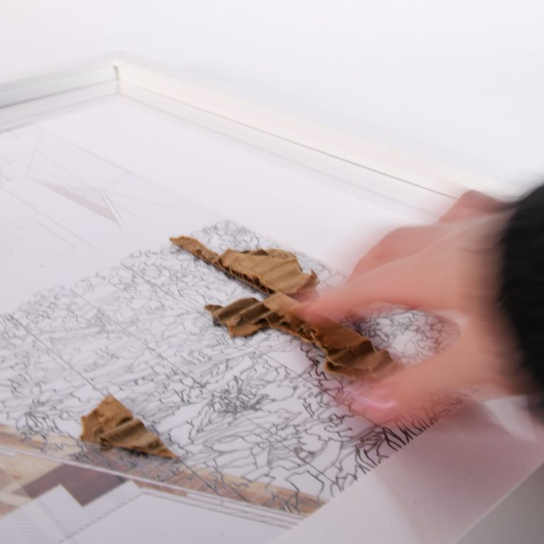 A hand arranges pieces of torn cardboard on top of a detailed, black-and-white line drawing placed on a flat surface.