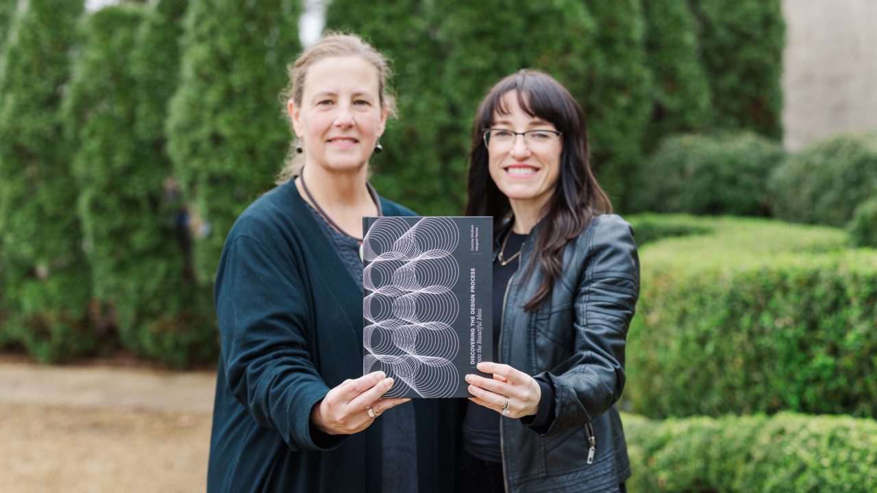 Two women standing outdoors hold up a black booklet with a geometric design on the cover, smiling at the camera; greenery is visible in the background.