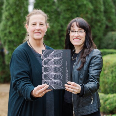 Two women standing outdoors hold up a black booklet with a geometric design on the cover, smiling at the camera; greenery is visible in the background.