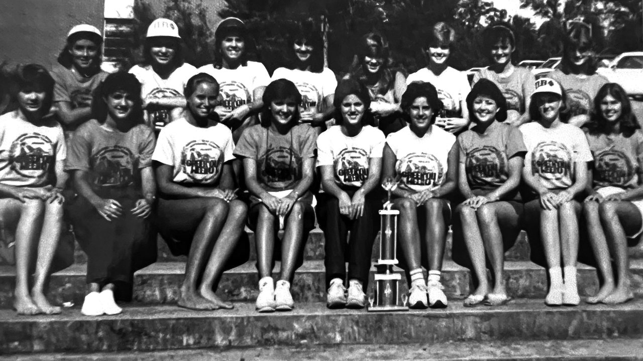 A group of sixteen young women pose on outdoor steps, some seated, some standing, all wearing matching t-shirts, with a trophy placed in front of the seated row.