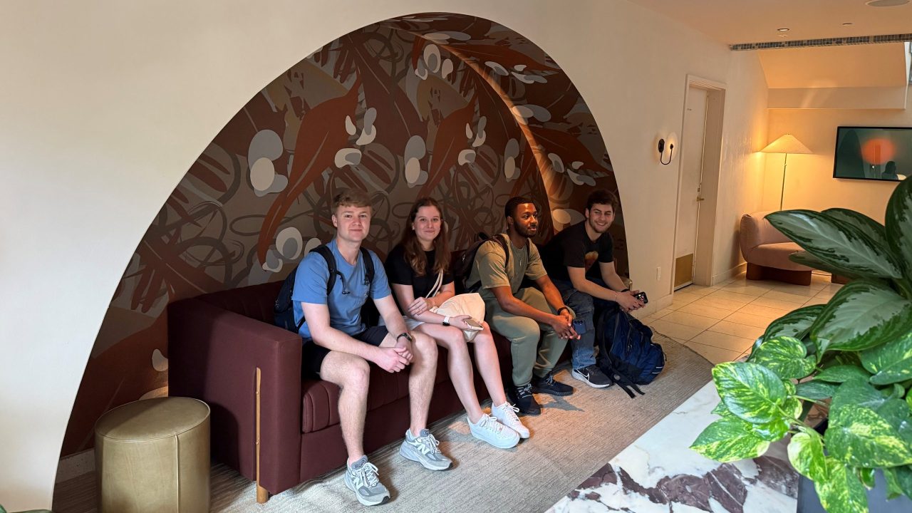 Four people with backpacks sit on a maroon couch under a decorative arch in a modern lobby, with plants and soft lighting nearby.