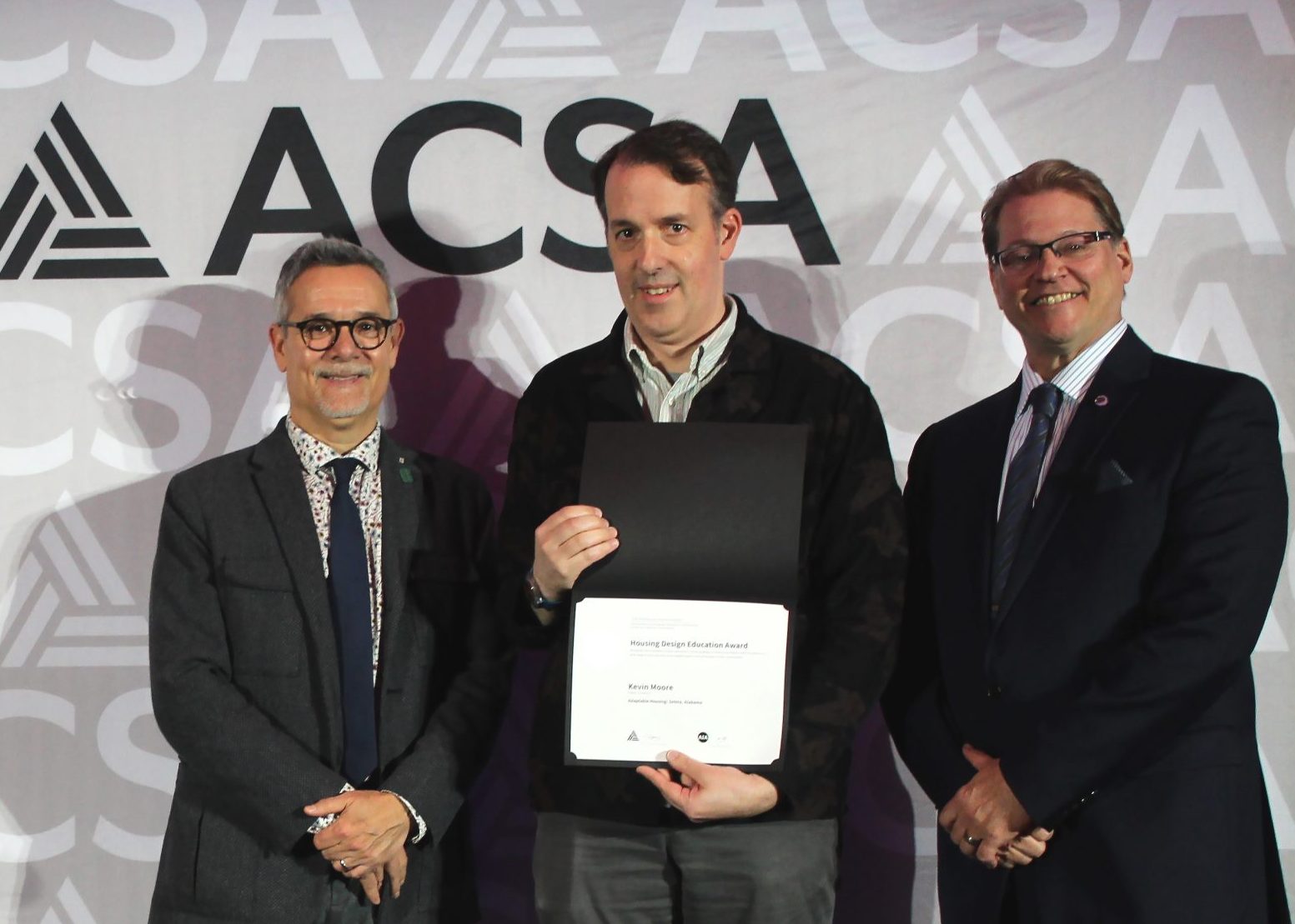 Three men stand in front of an ACSA backdrop; the man in the center holds a certificate and smiles at the camera.