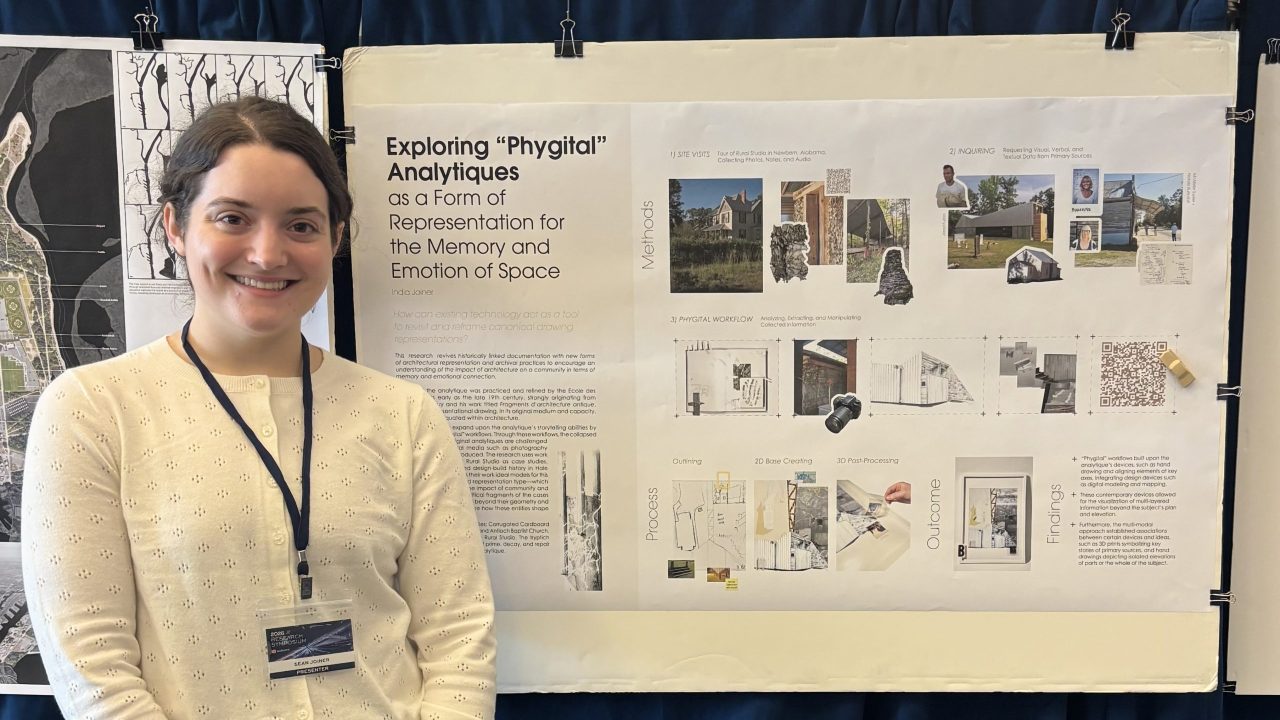 A woman stands beside a presentation board titled “Exploring ‘Phygital’ Analytiques,” which displays text, diagrams, and images related to memory and emotion in space.