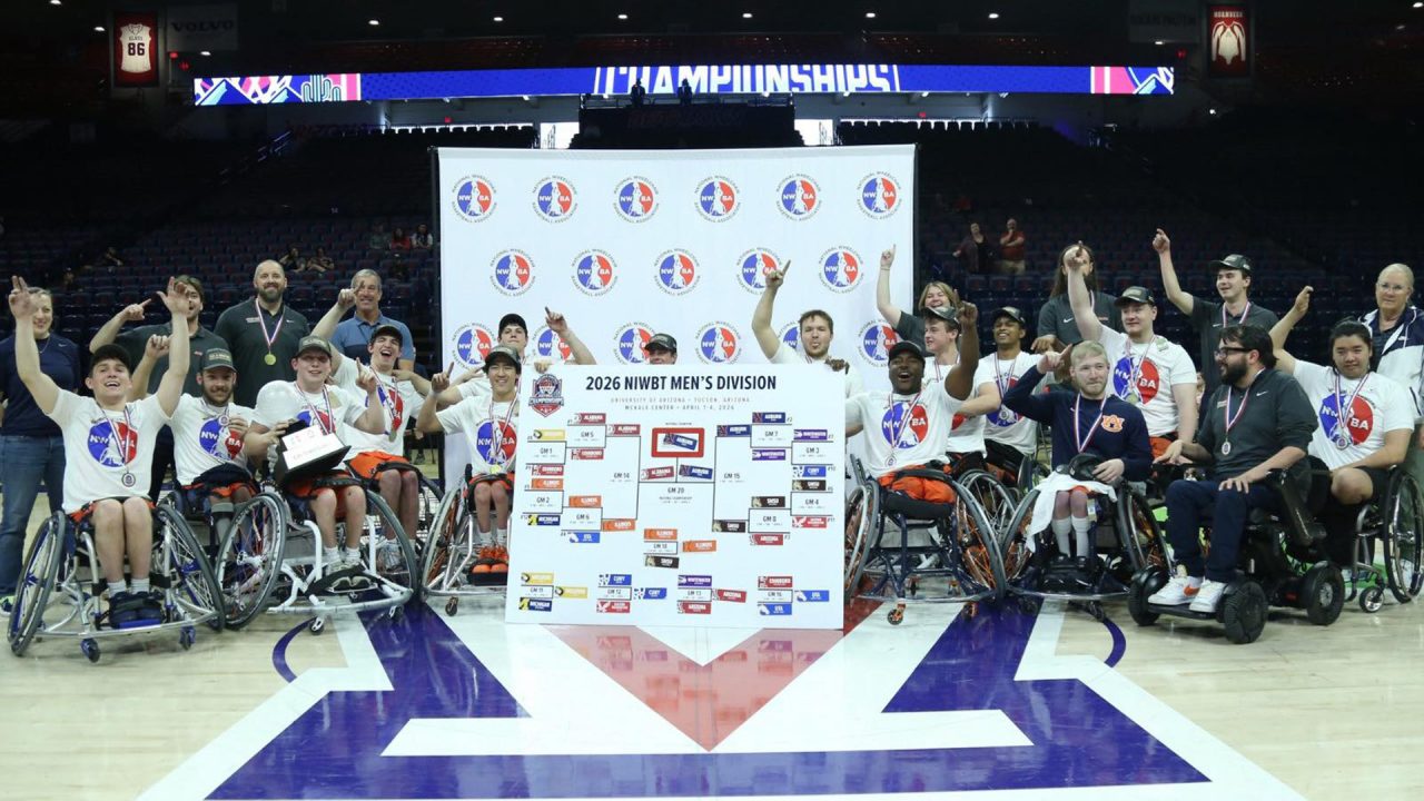 A wheelchair basketball team poses on a court with medals, shirts, and a large tournament bracket, celebrating their 2023 NWBT Men’s Division championship win.