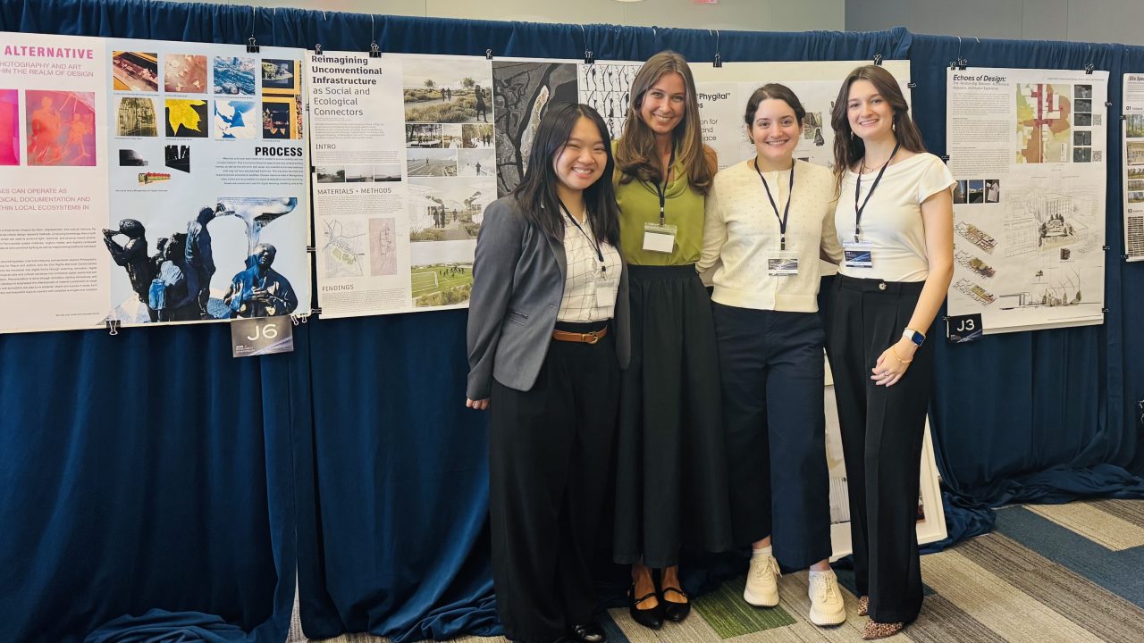 Four women stand and smile in front of a display of architectural posters at an indoor event, wearing conference badges.