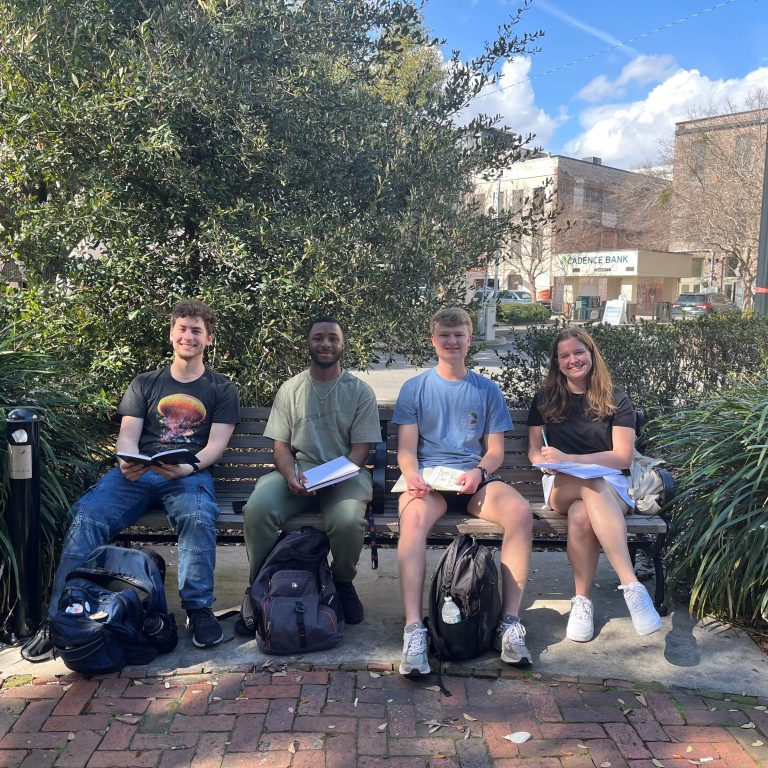 Four students sit on a bench outdoors, each holding papers and bags at their feet, with trees and buildings in the background.