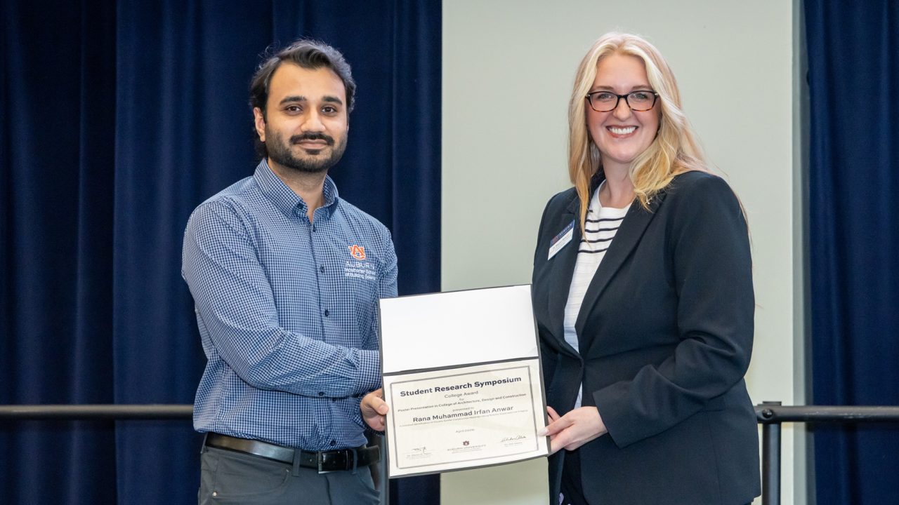 A man receives a certificate from a woman during a formal event, both smiling and standing in front of dark blue curtains.