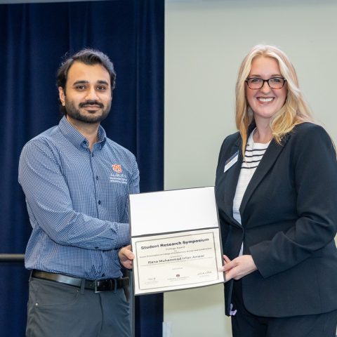 A man receives a certificate from a woman during a formal event, both smiling and standing in front of dark blue curtains.