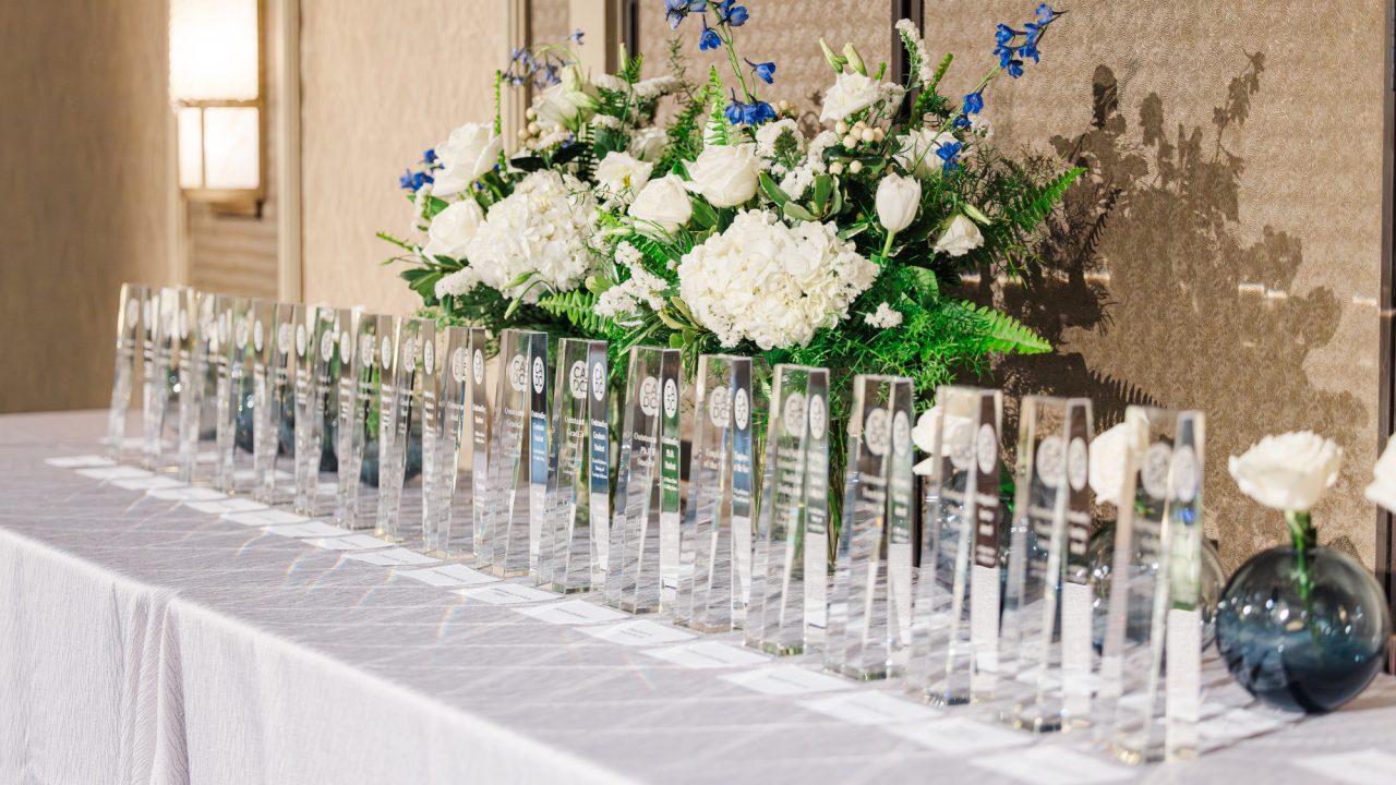 A row of clear glass awards is arranged on a white tablecloth, with a large floral arrangement of white and blue flowers in the background.