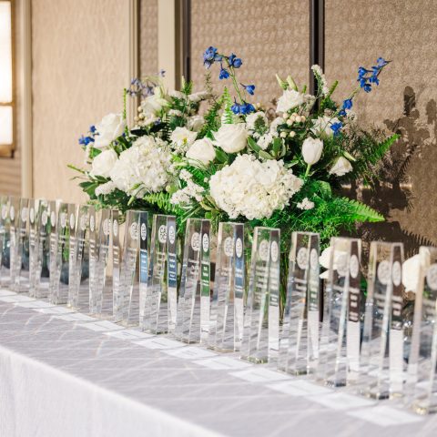 A row of clear glass awards is arranged on a white tablecloth, with a large floral arrangement of white and blue flowers in the background.