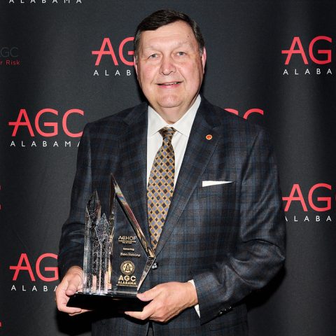 A man in a plaid suit holds an AGC Alabama award, standing in front of a backdrop with AGC Alabama and CompTrust logos.