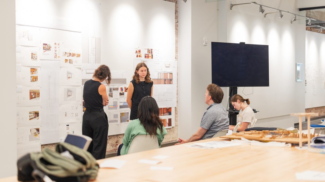A group of people are having a discussion in front of architectural drawings and plans displayed on a wall in a modern studio setting.