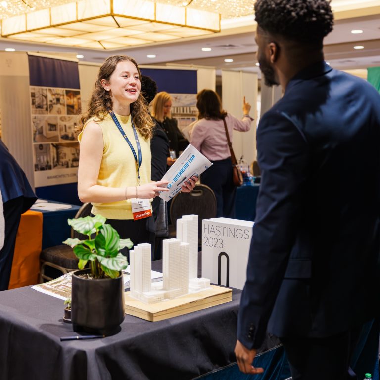 A woman stands at a booth with architectural models and talks to a man at an indoor event or career fair.