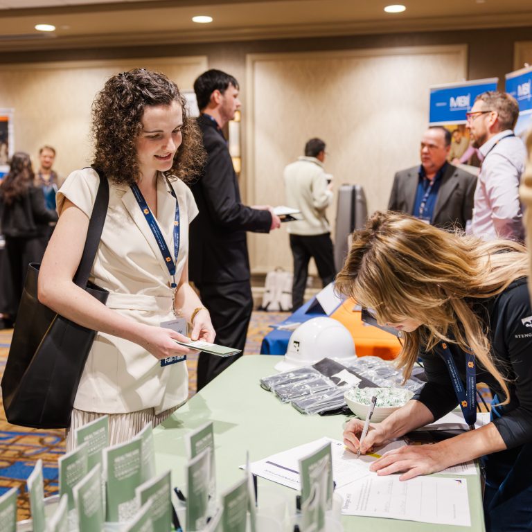 A woman stands at a registration table while another woman writes on a form at a business event; people are networking in the background.