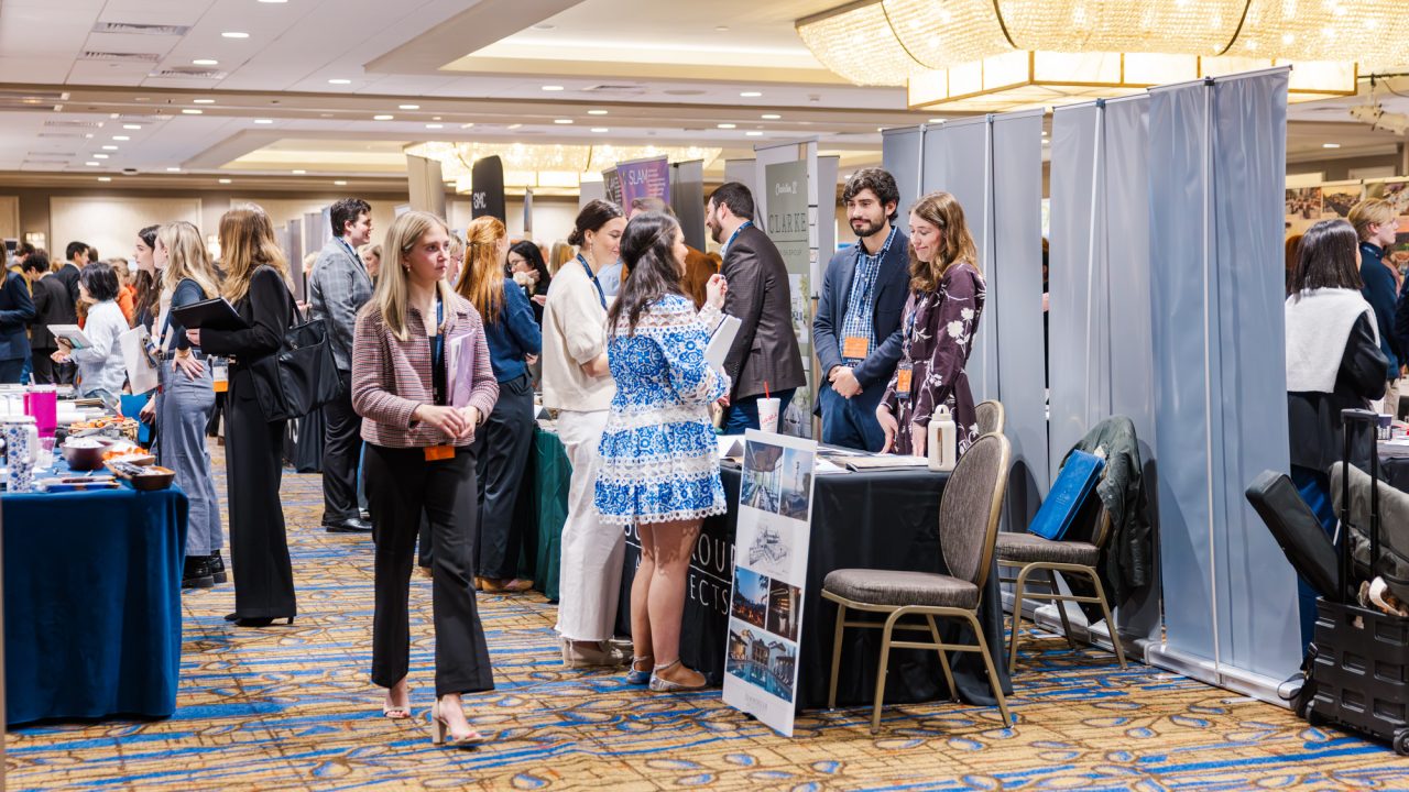 People attend a professional networking event or career fair in a large, well-lit conference room with booths and informational displays.