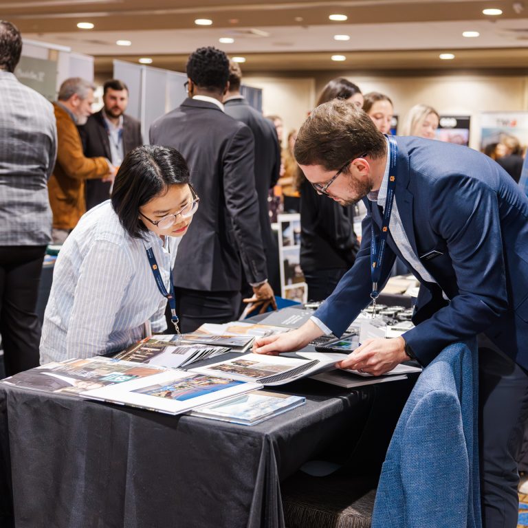 Two people review documents at a table during a busy business conference or networking event, with other attendees conversing in the background.