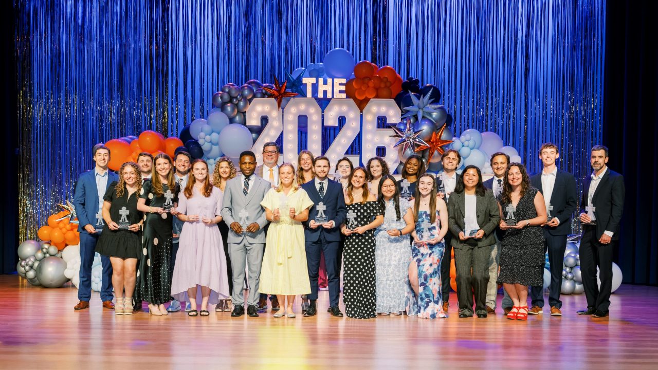 A group of people stands on stage holding trophies in front of a 2026 sign and balloon decorations.