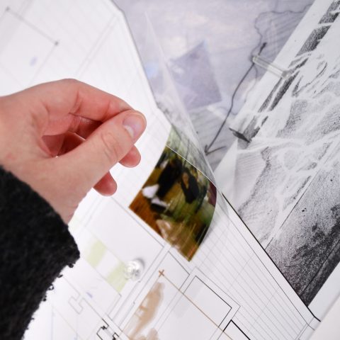 A hand lifts a transparent sheet over architectural plans, revealing a color photo underneath on a white background.