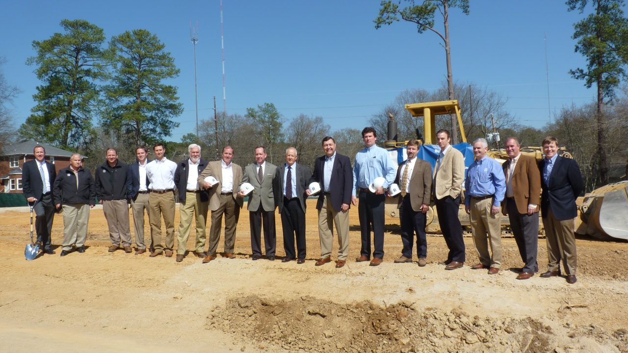 A group of men in business attire stand in a row at a construction site, holding hard hats and shovels, with construction equipment and trees in the background.