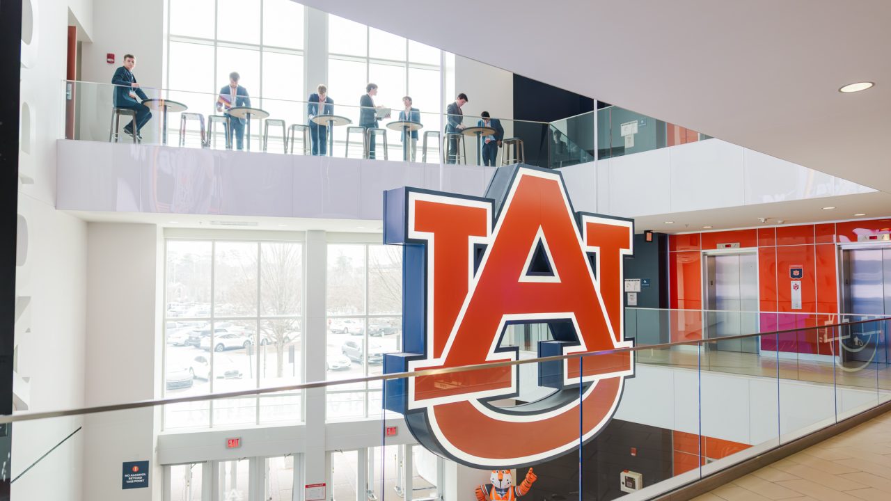 Large Auburn University logo displayed inside a modern building; several people sit at tables on an upper floor, and a person in a tiger mascot costume stands below.