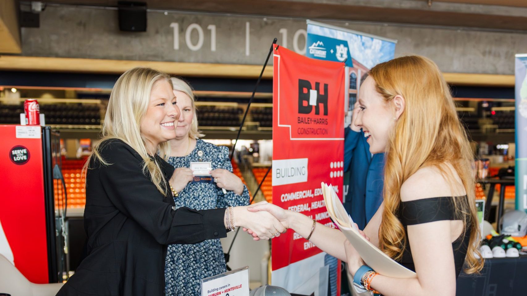 Two women stand behind a promotional table with Bailey Harris Construction branding, greeting a third woman at an indoor event.