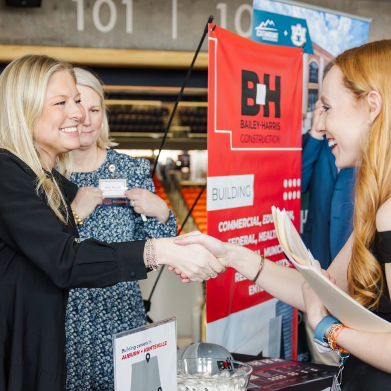 Two women stand behind a promotional table with Bailey Harris Construction branding, greeting a third woman at an indoor event.