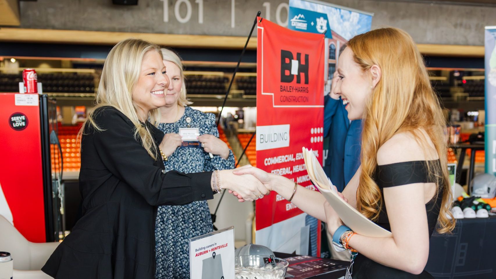 Two women stand behind a promotional table with Bailey Harris Construction branding, greeting a third woman at an indoor event.